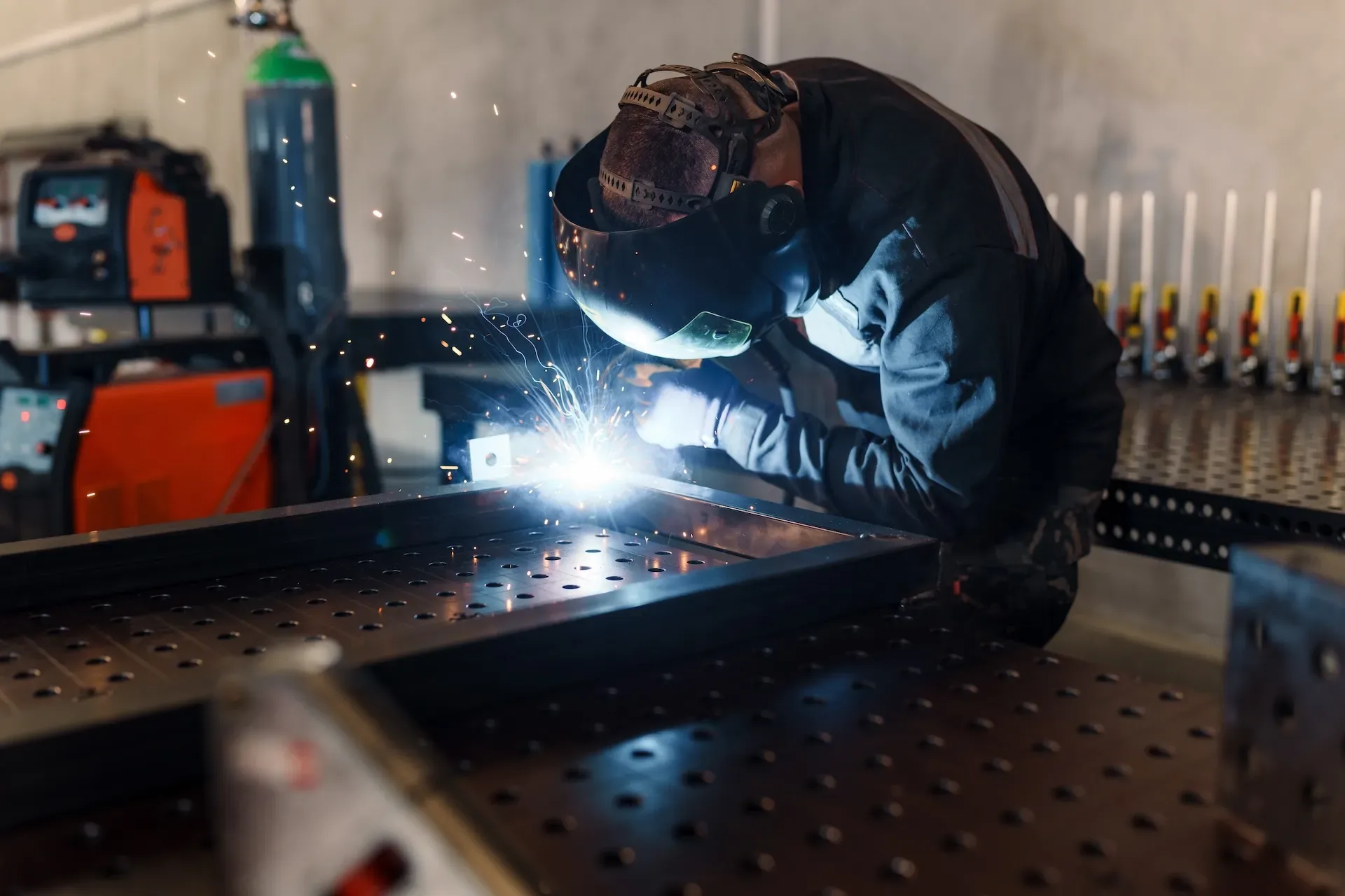 Welder in a helmet laying a bead on steel at a fabrication table, sparks flaring in a darkened workshop
